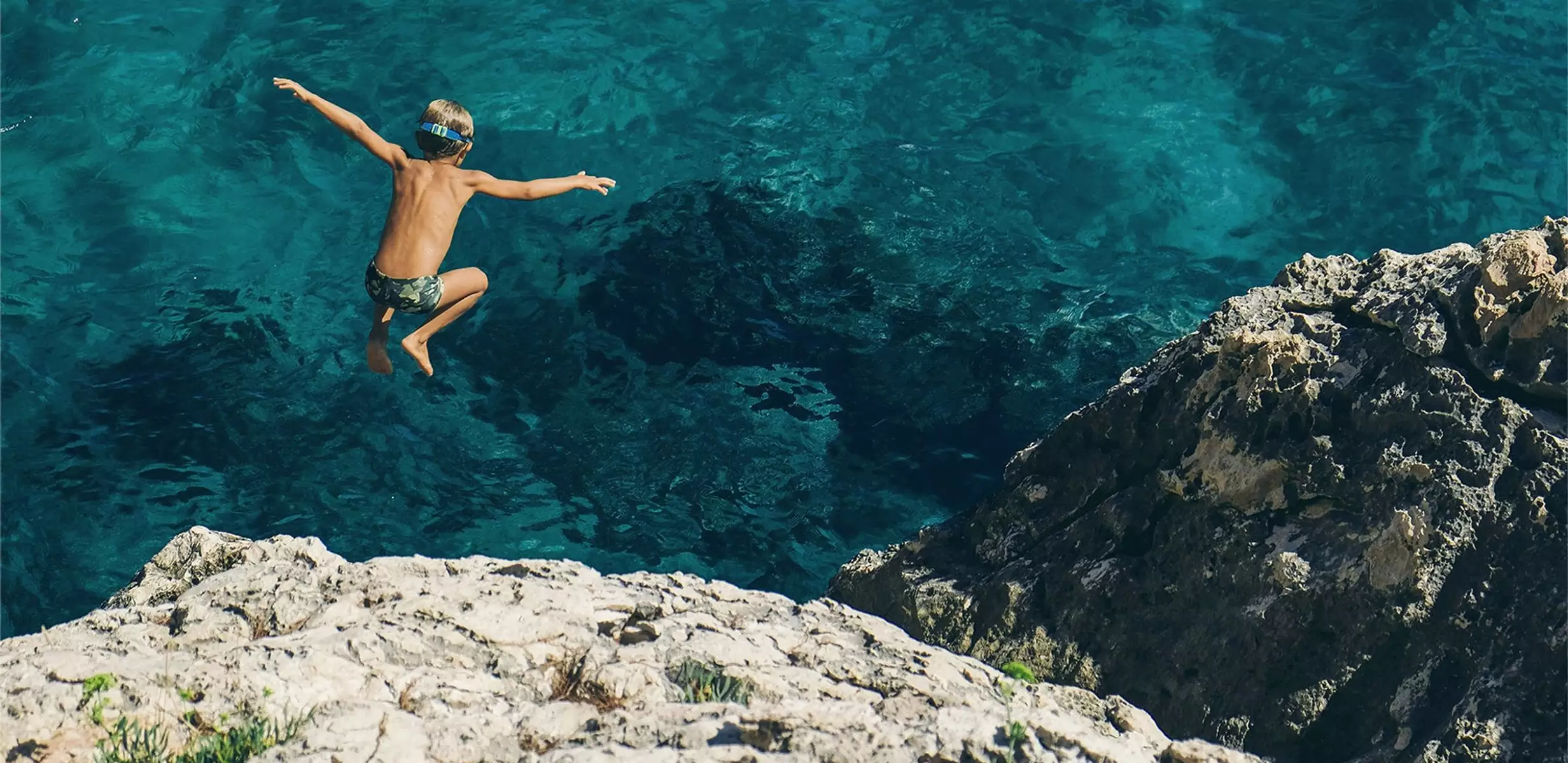 Niño saltando al mar desde unas rocas sobre aguas cristalinas de color turquesa en Menorca